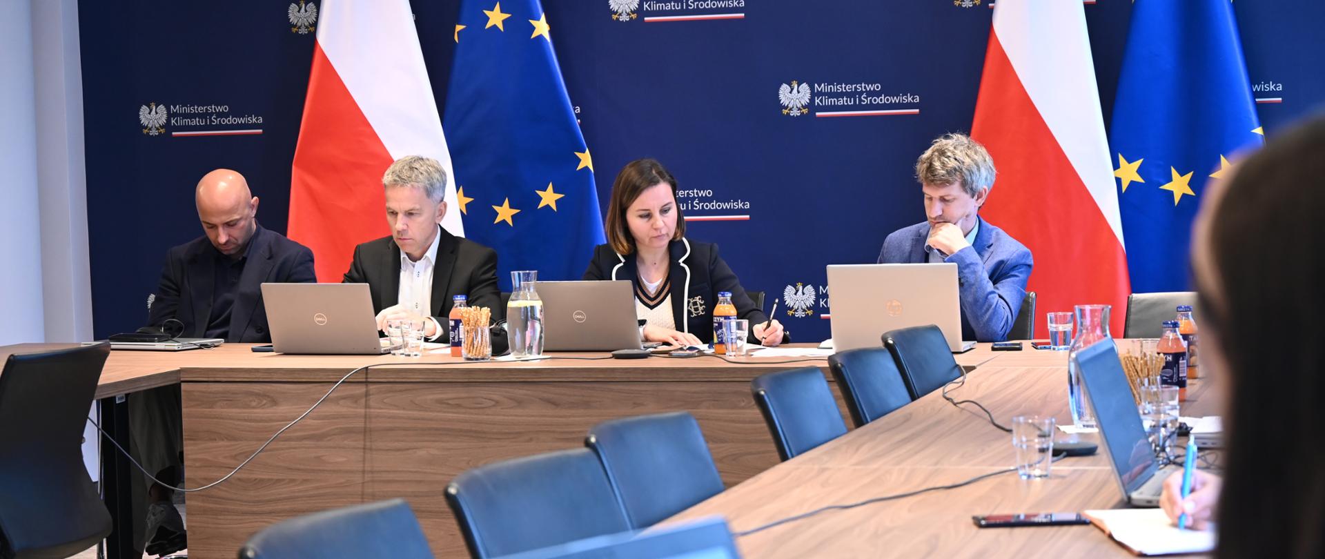 Cztery osoby siedzą z komputerami przy biurku. W centrum na tle flagi polskiej i EU siedzą członkowie Międzyresortowego Zespołu ds. Odry