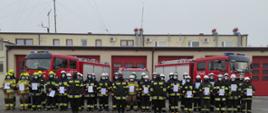 Zdjęcie ilustruje grupę strażaków ratowników OSP w umundurowaniu specjalnym, którzy ukończyli i odebrali zaświadczenia o ukończeniu szkolenia podstawowego. Strażacy stoją w dwuszeregu na tle dwóch pojazdów pożarniczych średnich GBA oraz jednego pojazdu gaśniczego lekkiego GLBA. Przed dwuszeregiem stoją trzej oficerowie KP PSP w Zduńskiej Woli w umundurowaniu koszarowym dowódczo-sztabowym, którzy wręczali zaświadczenia.
