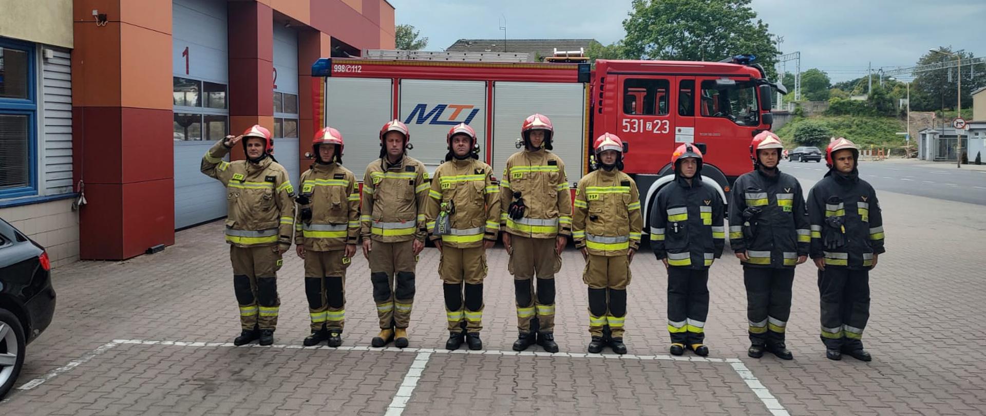 Pododdział strażaków na tle samochodu, dowódca salutuje.