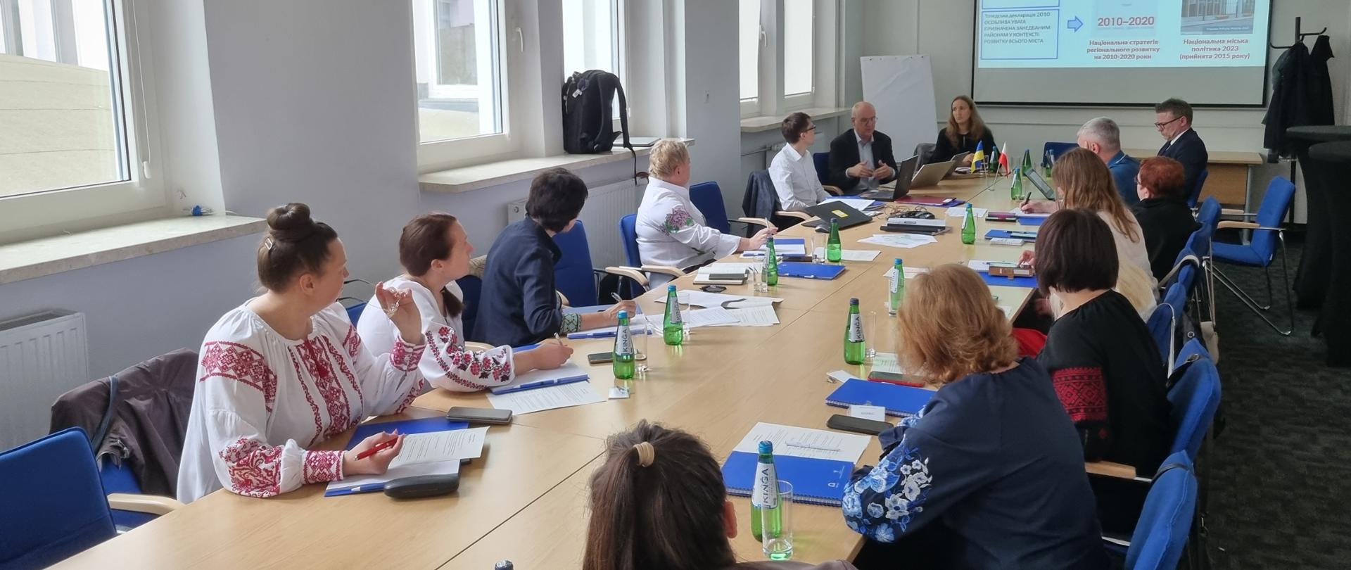Grupa osób siedzi przy długim stole konferencyjnym patrząc na prezentację. Część kobiet ubranych jest w tradycyjne koszule ukraińskie. Na stole leżą notatki