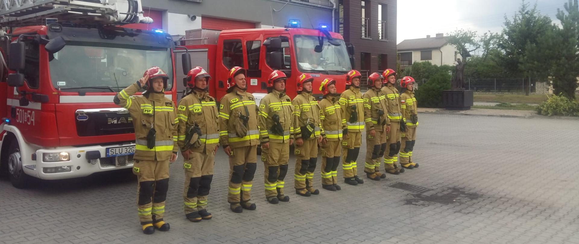 Na pierwszym planie strażacy ustawieni w szeregu, stojący na baczność. Oddają hołd w związku z Narodowym Świętem Dnia Pamięci Powstania Warszawskiego. Na drugim planie dwa samochody pożarnicze na tle budynku komendy.