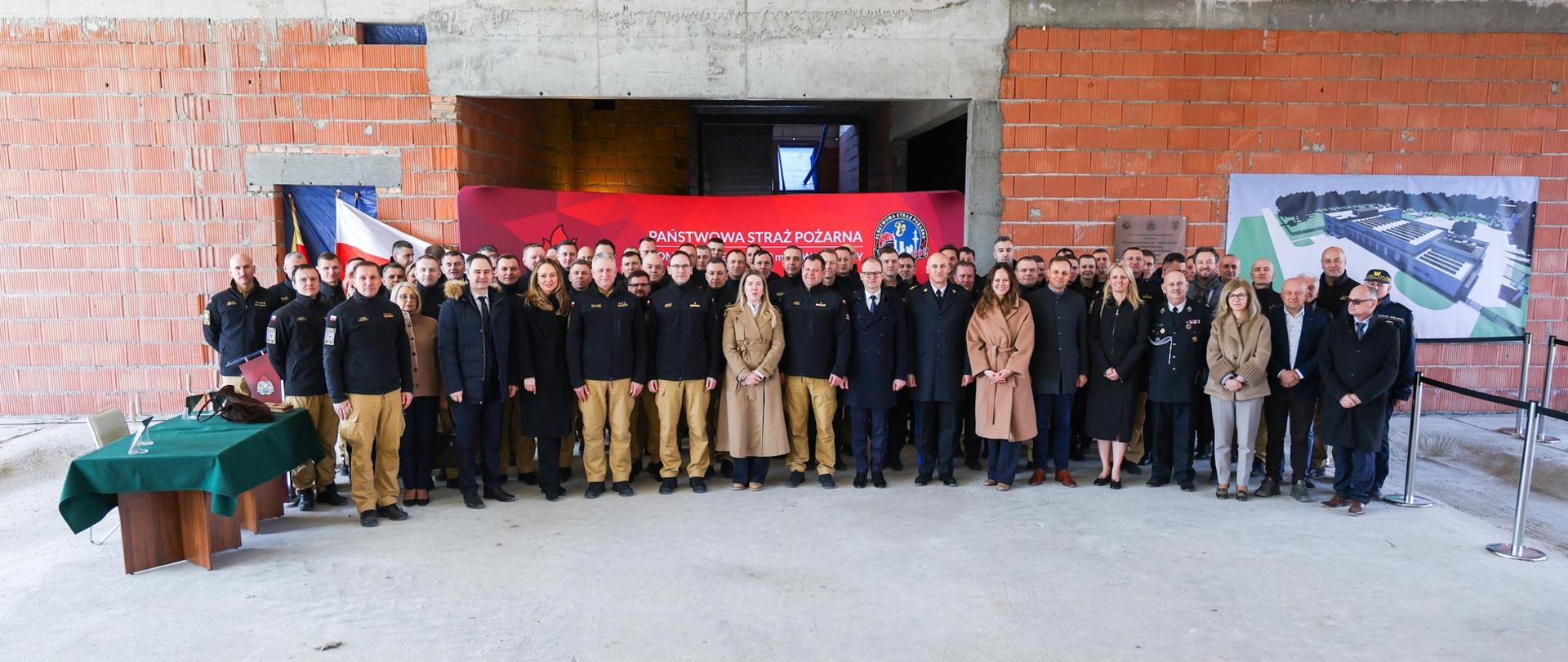 Zdjęcie grupowe wszystkich uczestników. Są wewnątrz budynku w stanie surowym z nieotynkowanymi ceglanymi ścianami. 