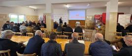 Sala wykładowa. W środkowej części osoba referująca. przed nim w ławkach siedzą ludzie.