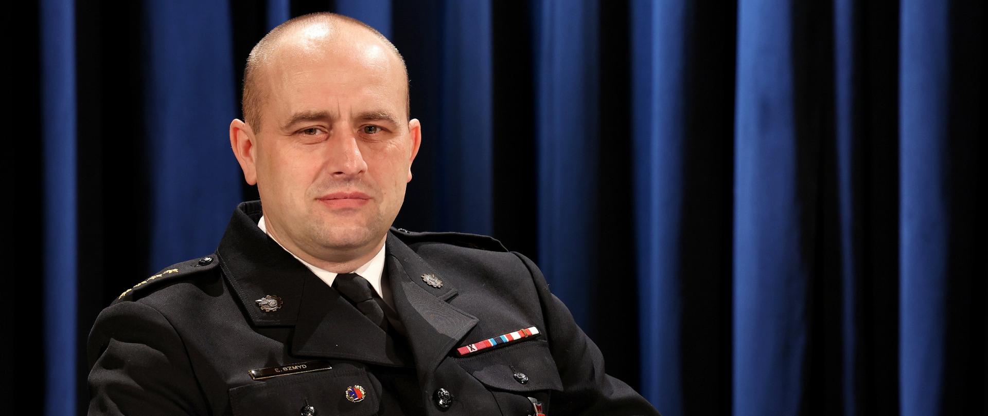 Zdjęcie przedstawia komendanta powiatowego PSP w Ustrzykach Dolnych st. kpt. mgr inż. Emila Szmyda który ubrany w mundur galowy z odznaczeniami siedzi na fotelu trzymając w rękach książkę. Komendant patrzy prosto w obiektyw a tło stanowi niebieska kotara.