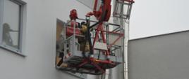 Ćwiczenia w GOBARTO. Zdjęcie przedstawia fragment budynku. Przy jednym z okien znajduje się kosz specjalnego samochodu z podnośnikiem hydraulicznym. W koszu znajduje się strażak, który podejmuje z pomieszczenia jedną z poszkodowanych osób. 
