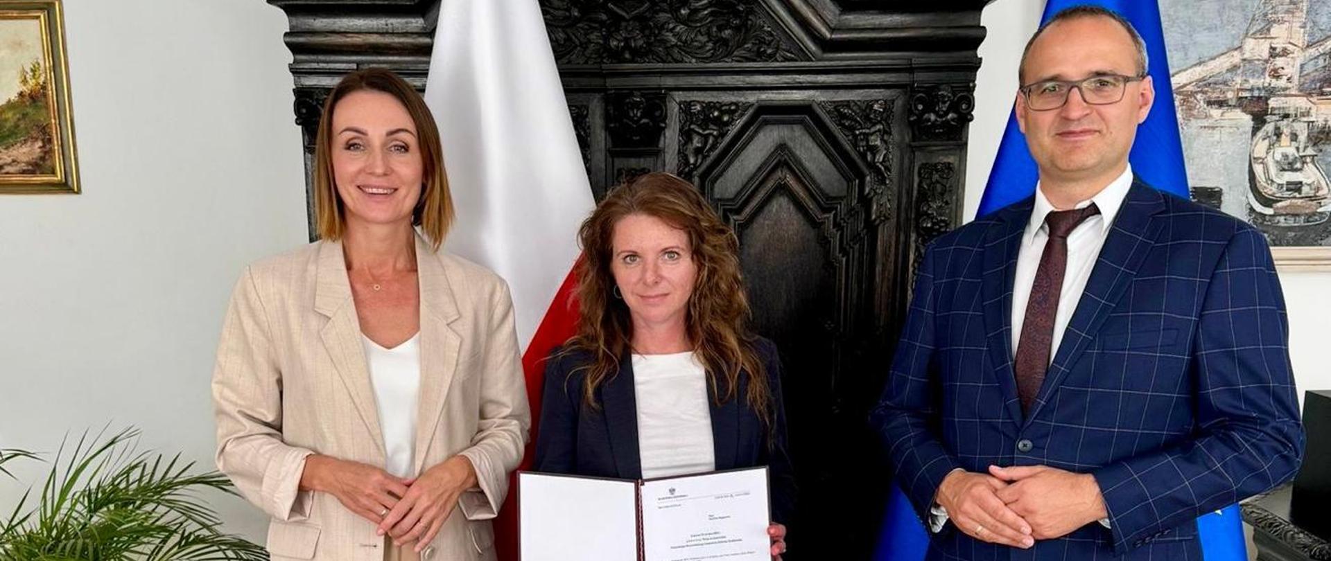 trzy osoby, dwie kobiety i mężczyzna stoją na tle flag polskiej i unijnej. Jedna z kobiet trzyma teczkę 