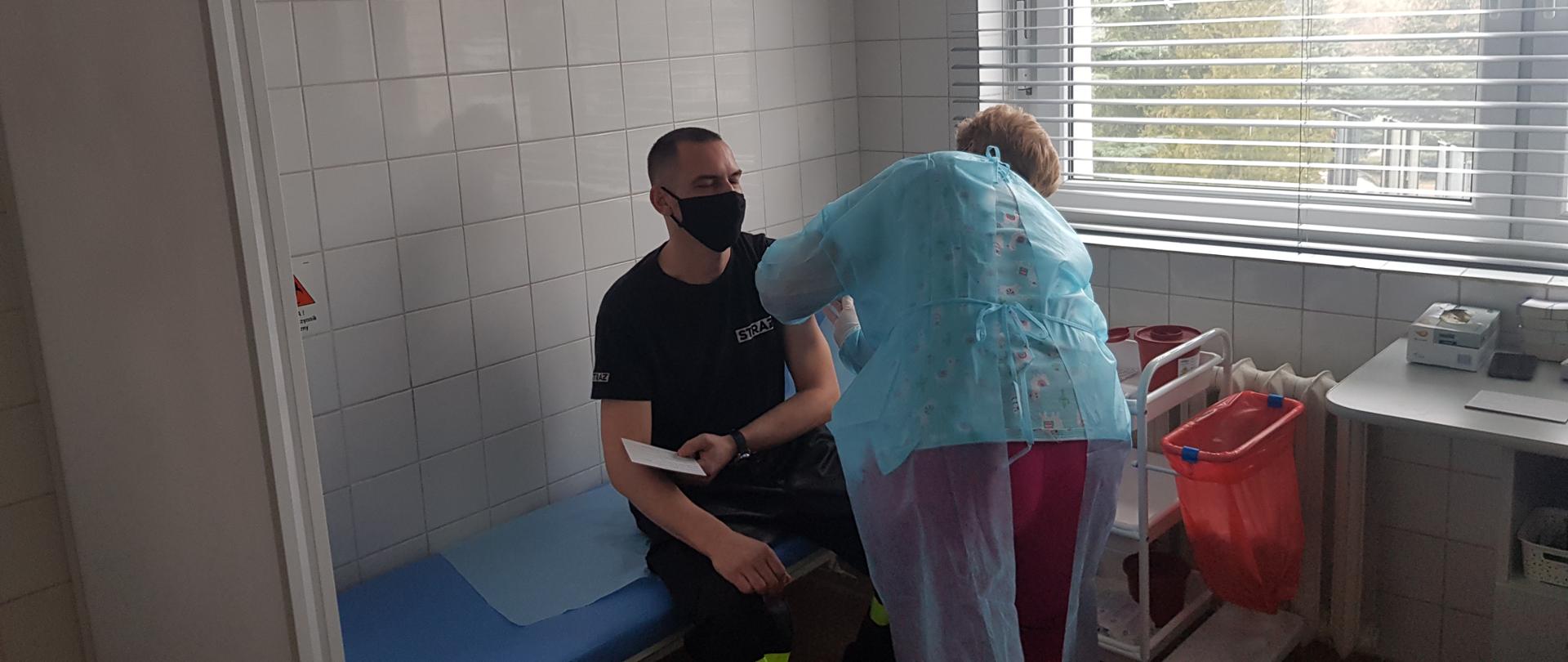 Jeden ze strażaków OSP siedzący na kozetce podczas szczepienia w gabinecie lekarskim przez Panią doktor.
