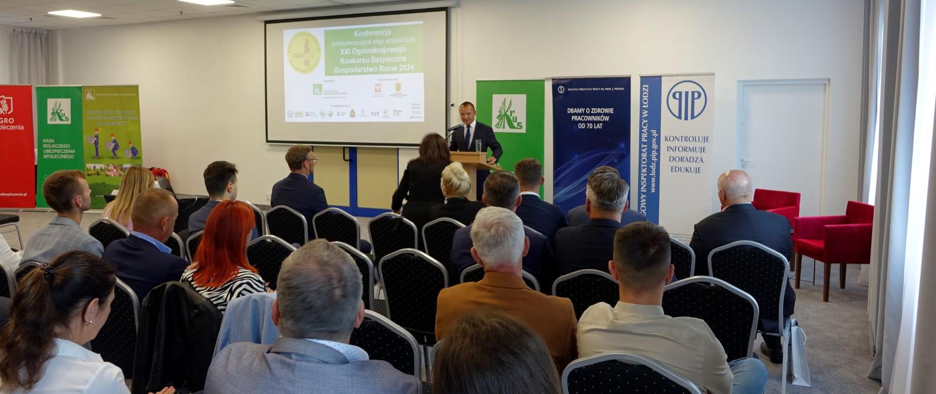 Mężczyzna przemawia przy pulpicie, obok ekranu z prezentacją z logo konkursu BGR, przed nim siedzi duża grupa osób na krzesłach.