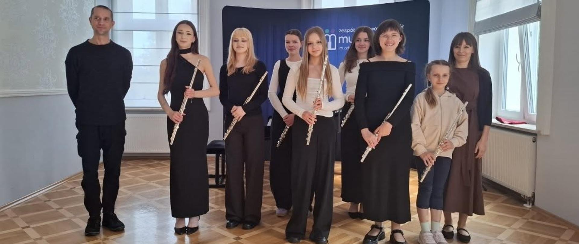 Zdjęcie przedstawia grupę dziewcząt – młodych flecistek – oraz dwoje dorosłych, pozujących do wspólnej fotografii w jasnej, eleganckiej sali koncertowej z tradycyjnym, drewnianym parkietem w kasetony.