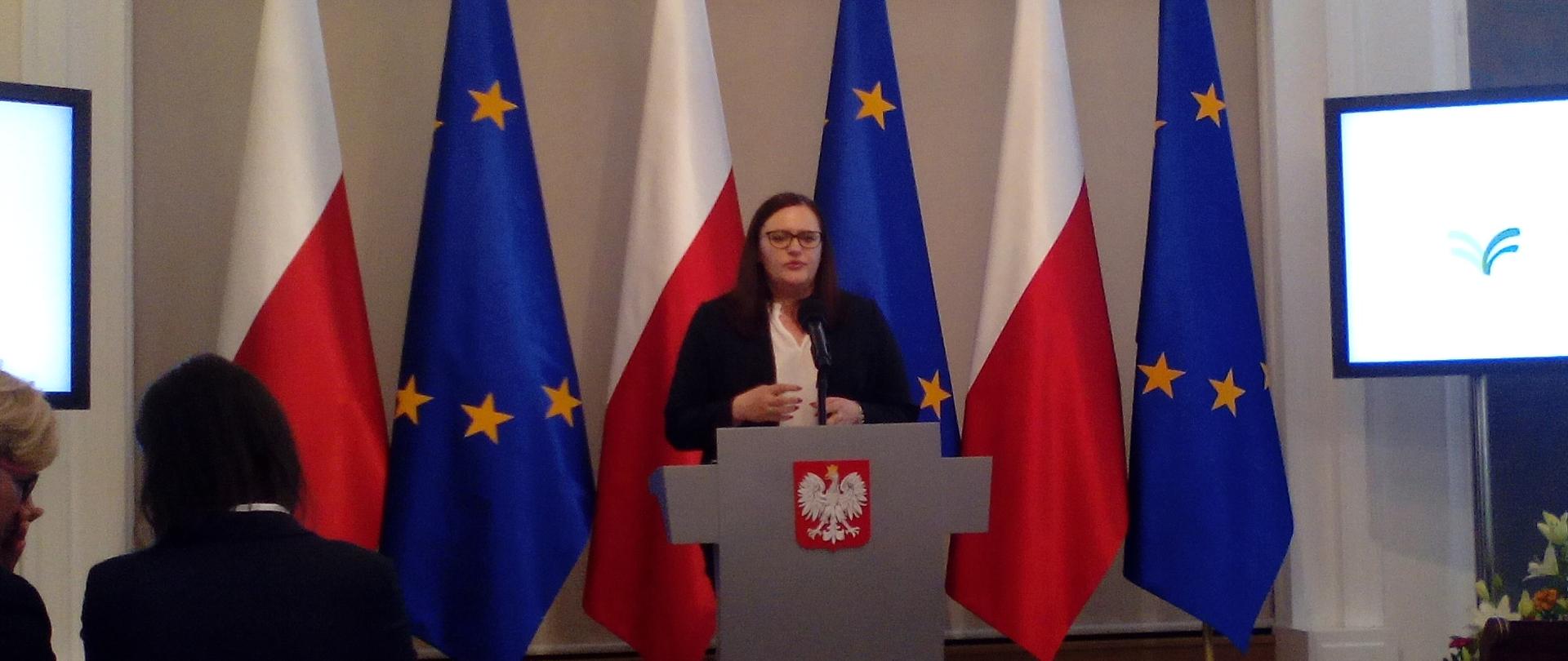 Minister Małgorzata Jarosińska-Jedynak przy mównicy, za nią sześć flag (trzy polskie i trzy unijne), po prawej i po lewej stronie ekrany z (niewidocznym na zdjęciu) logo wydarzenia i napisem "Perspektywy dla mieszkańców obszarów wiejskich". Z przodu widoczne kilka osób z widowni.