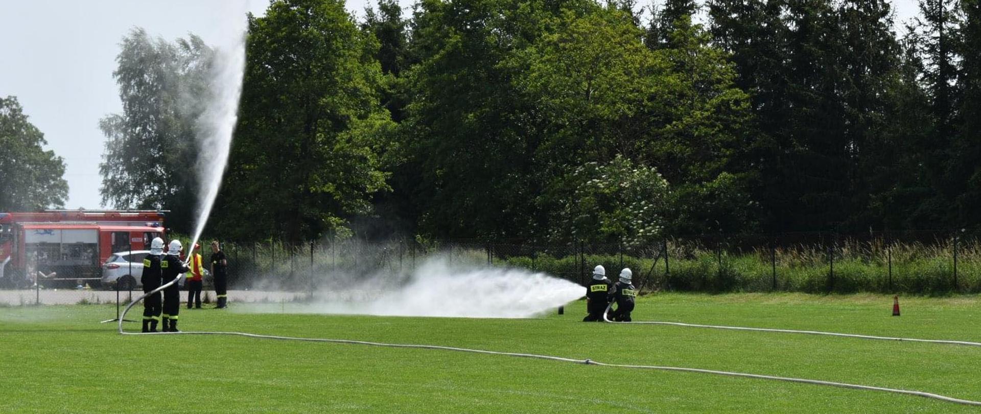 Czterech strażaków stojących po dwóch przy sobie, każda dwójka trzyma linię gaśniczą i celuje w przeszkody - pachołki i tarczę przed sobą. Strażacy w czarnych ubraniach koszarowych i białych hełmach. Jest to rozwinięcie w ramach ćwiczenia bojowego na murawie boiska.