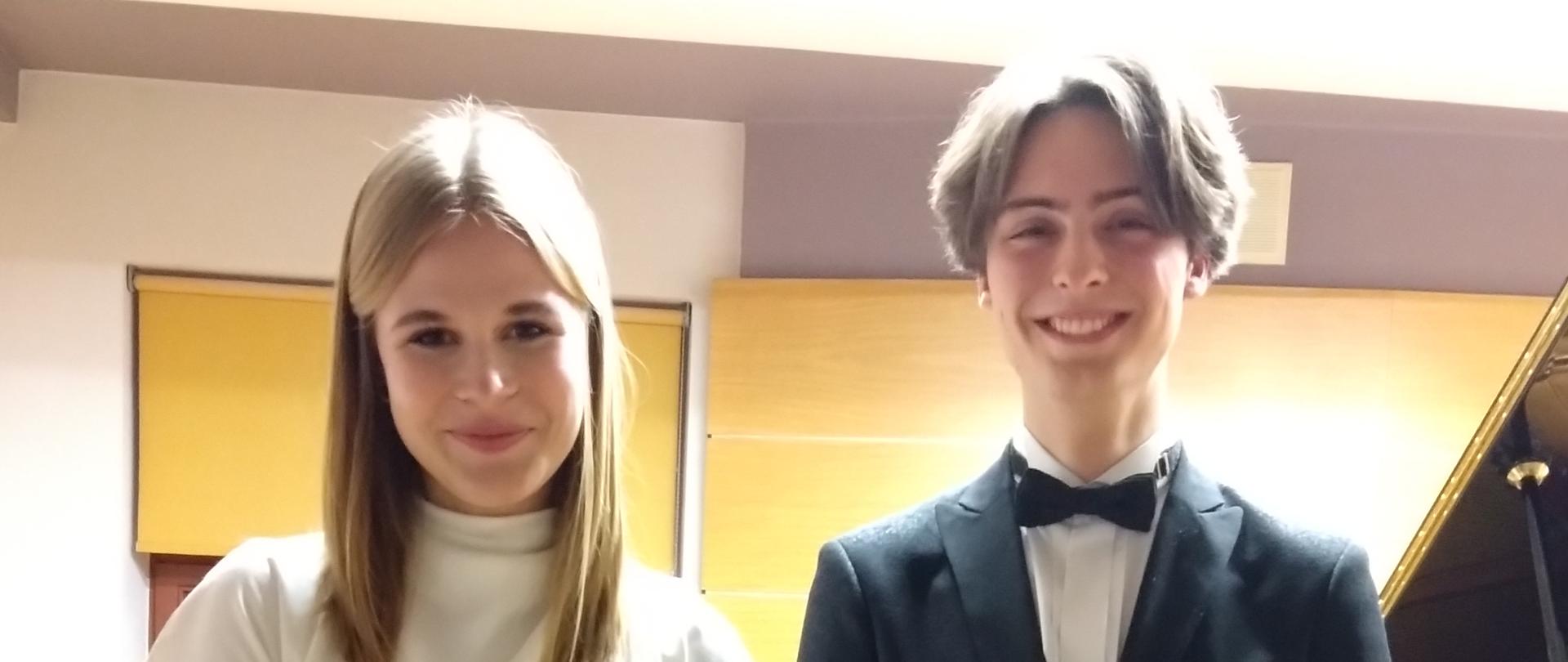 Uczniowie grający audycje stoją na scenie sali kameralnej. Za nimi dwa fortepiany raz ławy fortepianowe. W tle okno przysłonięte roletą. Pod oknem nawilżacz powietrza oraz grzejnik