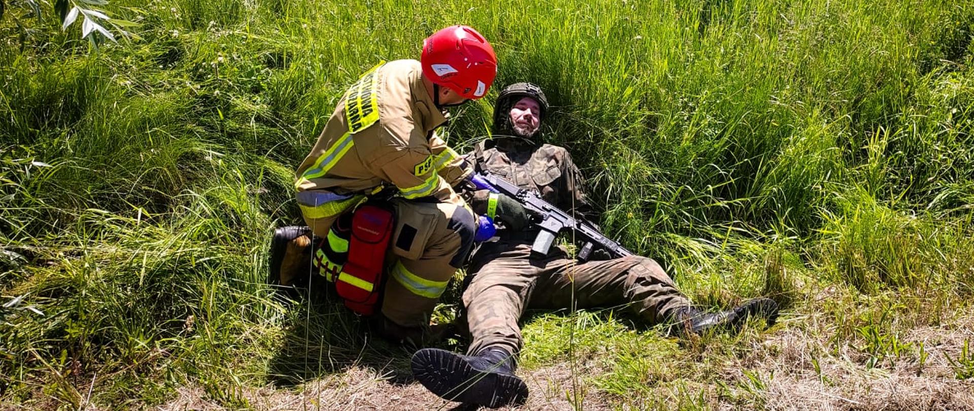 Strażaka, który prowadzi segregację (triage). Przyczepia on opaskę leżącemu żołnierzowi. Żołnierz leży na trawie i trzyma w ręku karabin.
