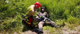 Strażaka, który prowadzi segregację (triage). Przyczepia on opaskę leżącemu żołnierzowi. Żołnierz leży na trawie i trzyma w ręku karabin.