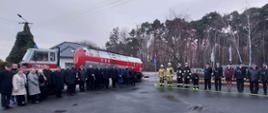 Strażacy w mundurach i osoby cywilne stojąna tle samochodu cysterny na placu.
