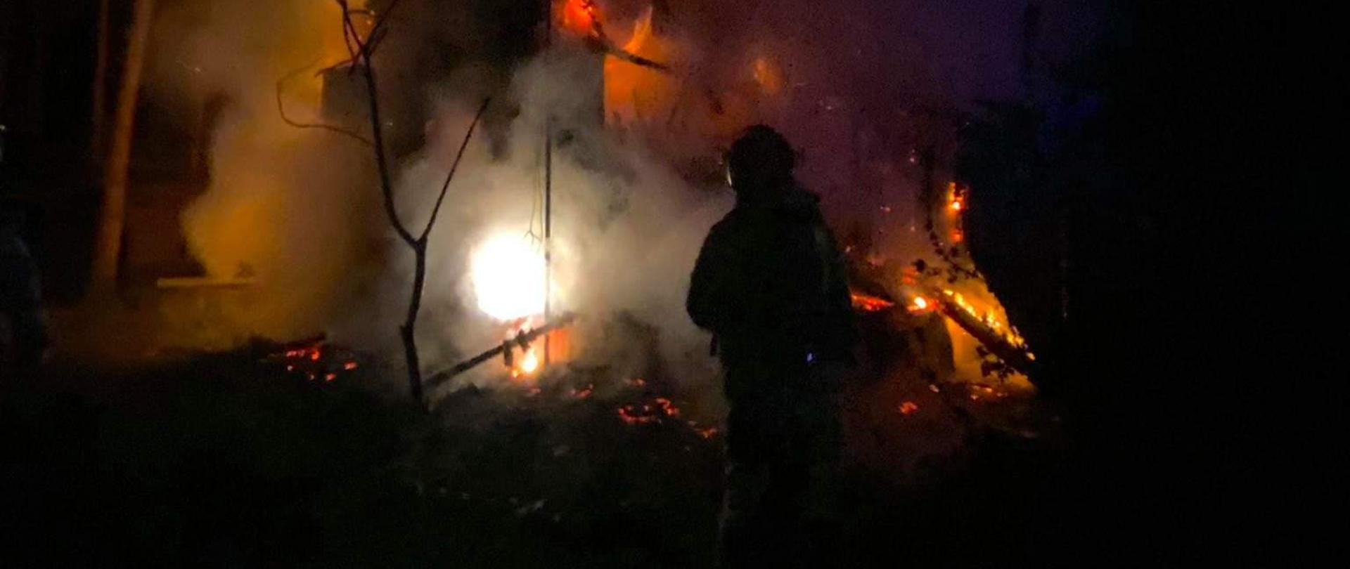 Na środku zdjęcia widoczny jest drewniany budynek w całości objęty pożarem. Płomienie wydostają się poprzez przepalony dach oraz okna , a także zniszczone przez ogień ściany budynku. Przed płonącym budynkiem widoczny jest strażak który podawał prąd wody na palący się budynek. Na lewo od strażaka widać stojące cienkie drzewo , a wokół niego dopalające się elementy budynku. W oddali po lewej stronie zdjęcia widać drzewa , które znajdują się za budynkiem.