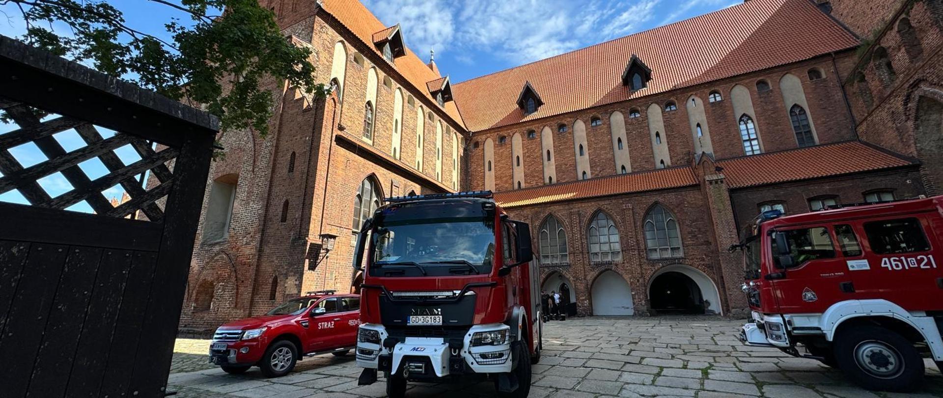 Na dziedzińcu Zamku w Kwidzynie stoją dwa samochody gaśnicze oraz jeden lekki samochód operacyjny.