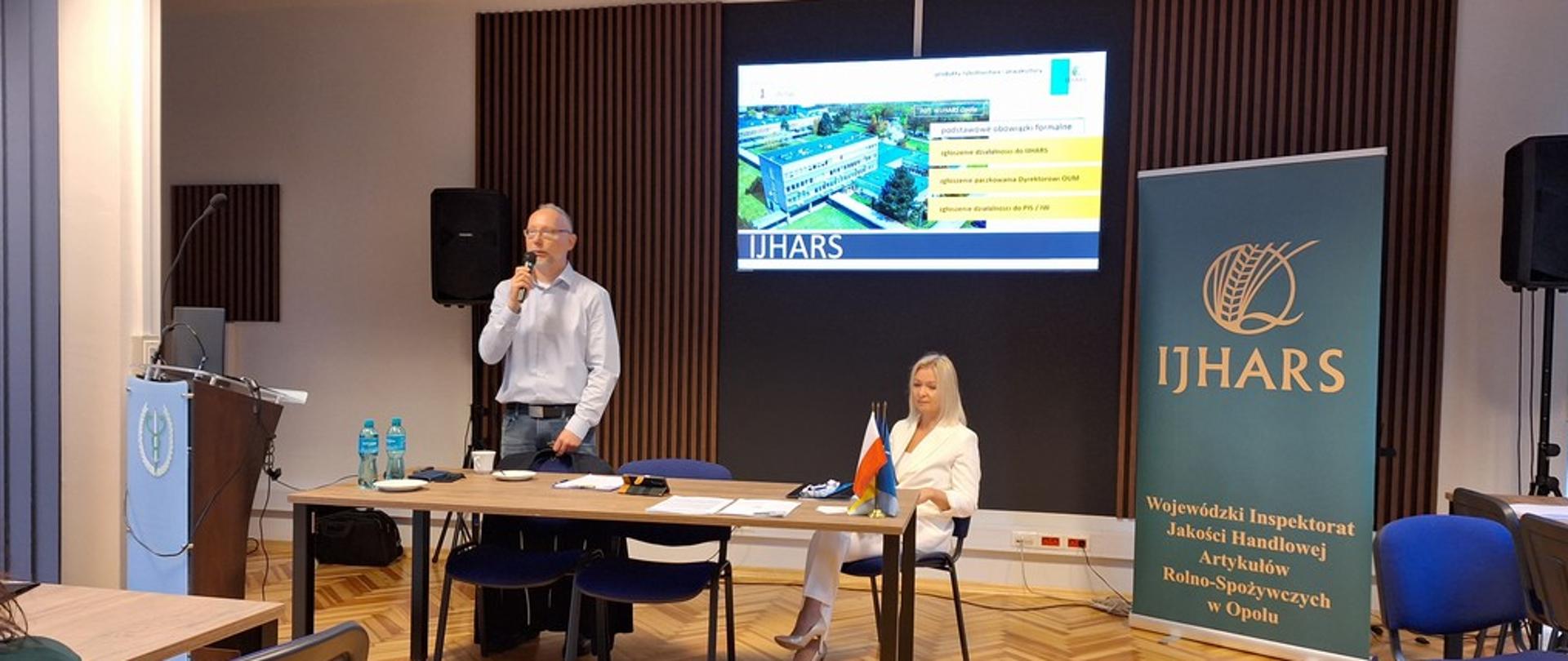 Przy stoliku stoi prowadzący szkolenie. W prawej dłoni, przy ustach mikrofon. Obok siedzi p. Dorota Szyra - Opolski Wojewódzki Inspektor Jakości Handlowej Artykułów Rono-Spożywczych. W tle banner z logo i napisem IJHARS, Z tyłu wyświetlony slajd z fotografią siedziby Inspektoratu