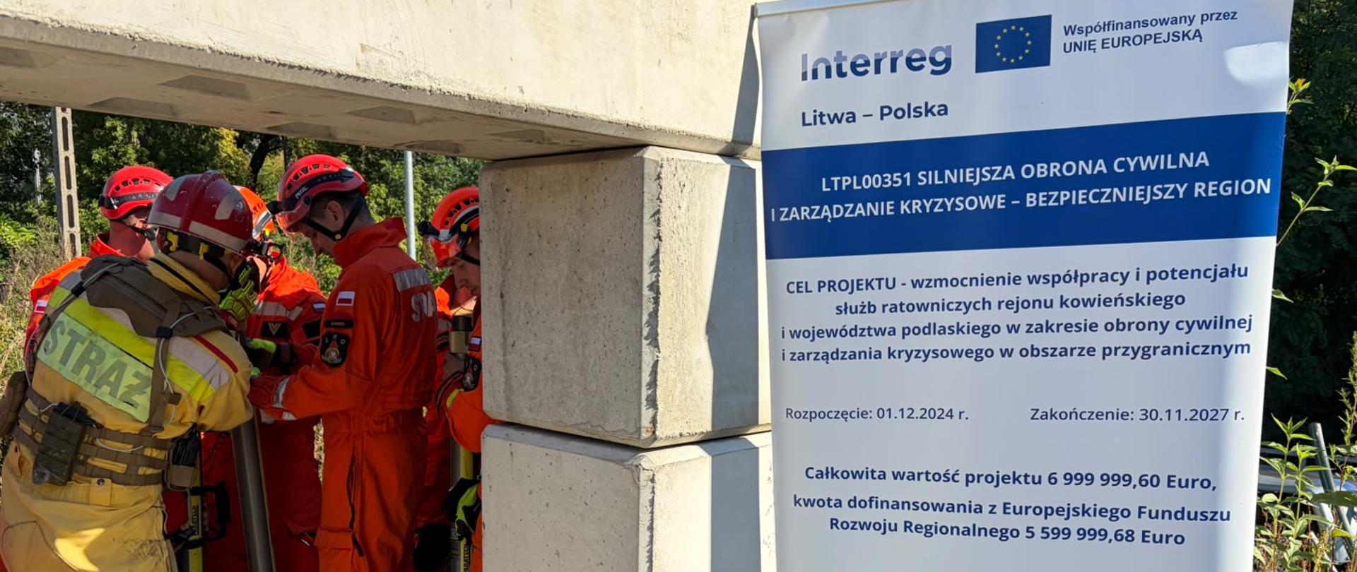 Zakończyło się polsko-litewskie szkolenie grup poszukiwawczo-ratowniczych - strażacy z lewej strony stoją - mają na sobie ubrania specjalne w kolorze pomarańczowym i żółtym, a także kaski i specjalne obuwie, po ptawej stronie stoi rollup promujący program Interreg