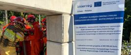 Zakończyło się polsko-litewskie szkolenie grup poszukiwawczo-ratowniczych - strażacy z lewej strony stoją - mają na sobie ubrania specjalne w kolorze pomarańczowym i żółtym, a także kaski i specjalne obuwie, po ptawej stronie stoi rollup promujący program Interreg