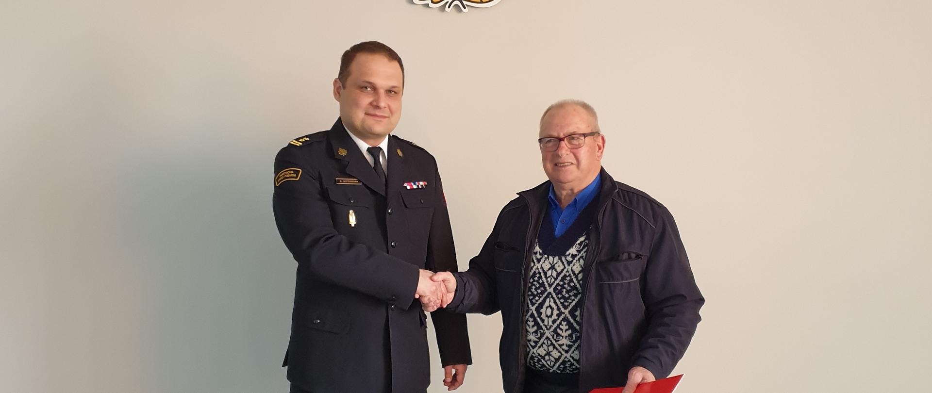 Na zdjęciu Komendant Miejski Państwowej Straży Pożarnej bryg. Sławomir Kotoński w ubraniu wyjściowym PSP i druh. Zdjęcie w pomieszczeniu. Na podłodze szary dywan. W tle herb oraz logo.