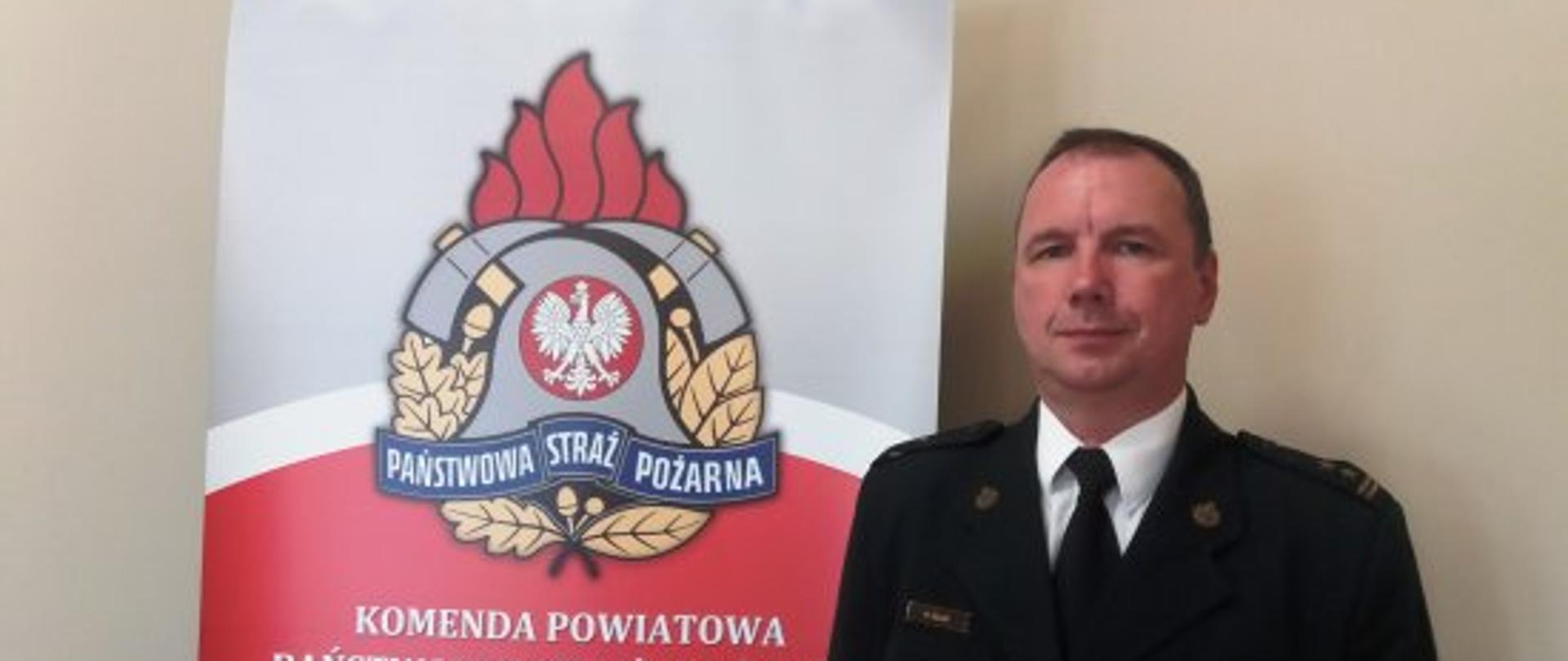 na zdjęciu widoczny jest strażak w umundurowaniu wyjściowym, w ciemnej marynarce z dystynkcjami, białej koszuli z czarnym krawatem. Strażak stoi na tle jasnej ściany obok baneru z logiem państwowej straży pożarnej i napisem komenda powiatowa państwowej straży pożarnej w krotoszynie