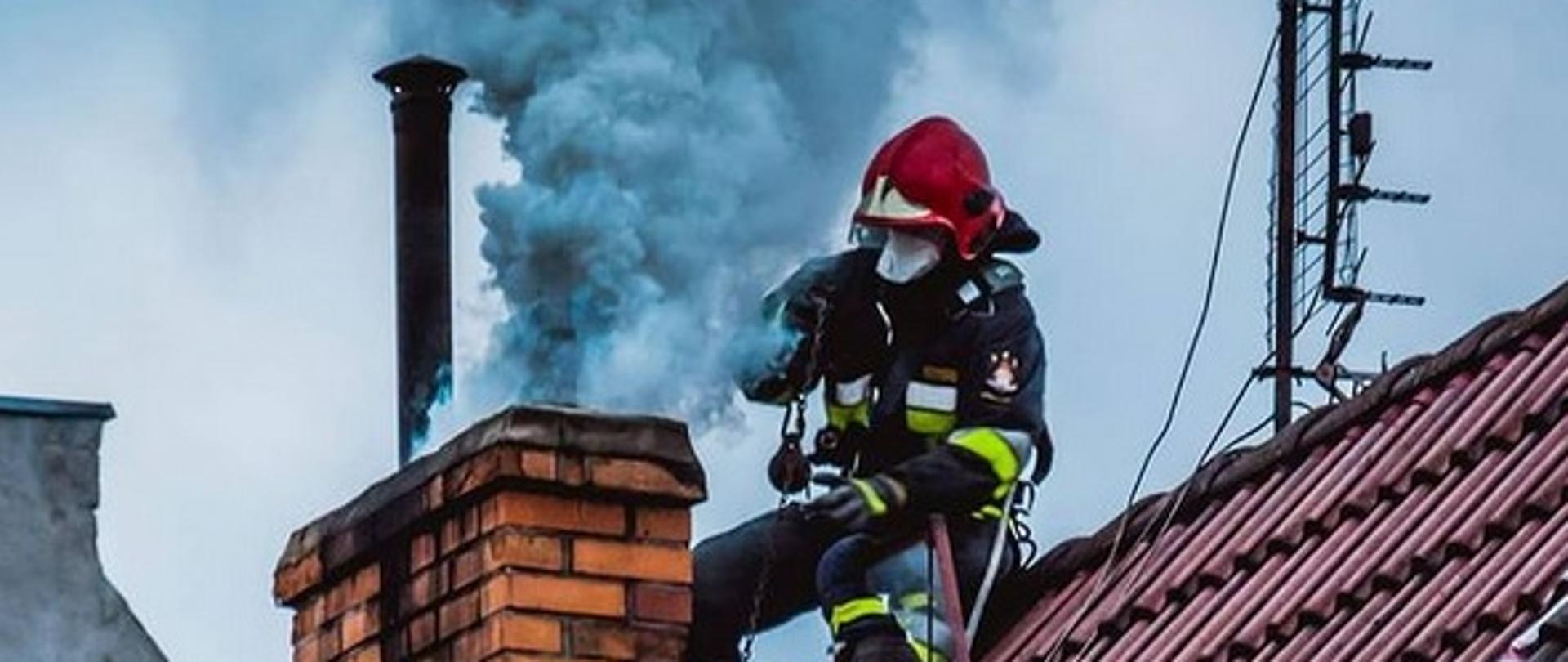 Zdjęcie przedstawia dym wydobywający się z komina podczas pożaru przewodu kominowego. Na dachu przy kominie znajdują się strażacy gaszący pożar. 