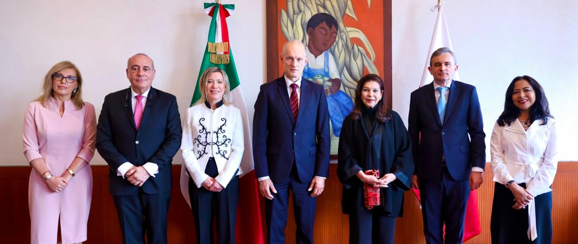 Siedem osób stoi w rzędzie na tle flag. Uczestnicy są ubrani formalnie - w garnitury i eleganckie stroje. 