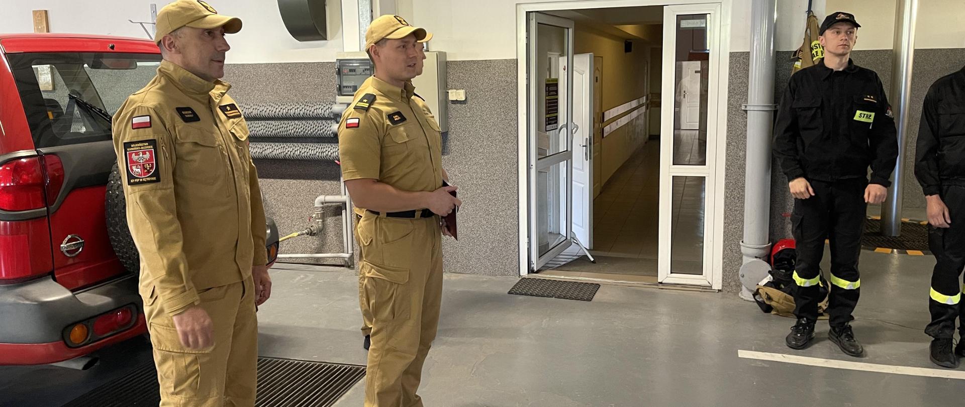 Po lewej stronie można zauważyć Komendanta Powiatowego Państwowej Straży Pożarnej w Kętrzynie oraz Zastępcę Komendanta Powiatowego Państwowej Straży Pożarnej w Kętrzynie ubranych w żółte stroje służbowe. Za nimi stoi czerwony samochód strażacki. Po lewej stronie stoją strażacy w czarnych strojach koszarowych. 