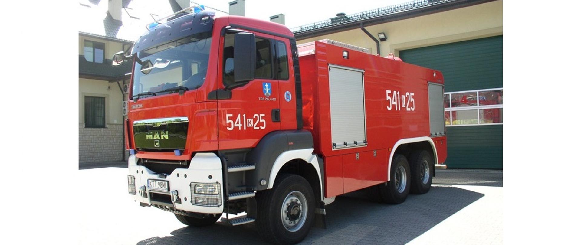 Zdjęcie przedstawia ciężki samochód z zakopiańskiej Jednostki ratowniczo gaśniczej w Zakopanem. W tle Budynek Komendy Powiatowej PSP w Zakopanem. Zdjęcie zostało wykonane w słoneczny dzień.