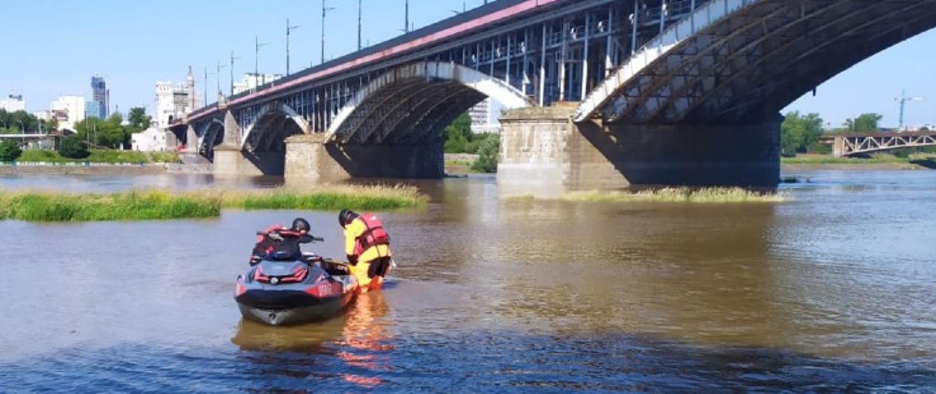 Na fotografii widać skuter wodny unoszący się na wodzie tuż przy brzegu. Obok pojazdu stoją dwaj strażacy ubrani w skafandry do ratownictwa wodnego. Ratownicy montują specjalną pływająca platformę do skutera. Na drugim planie widać drugi brzeg rzeki. Nad całością góruję Most Śląsko-Dąbrowski.