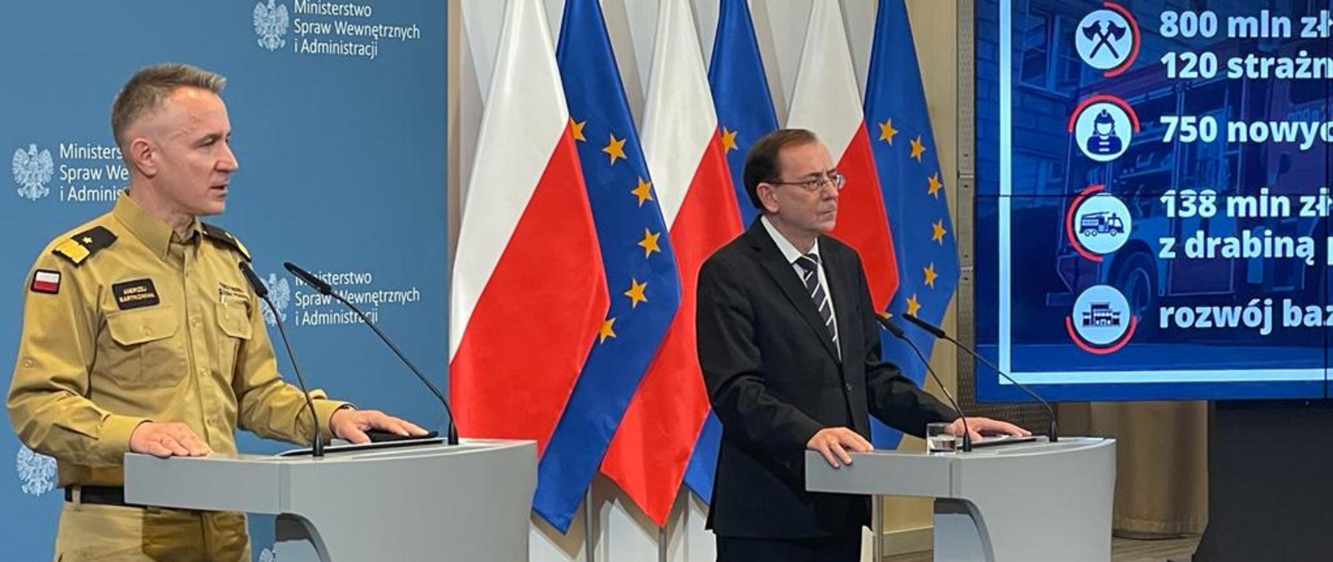 Stojący przy mównicach Komendant Główny Państwowej Straży Pożarnej oraz minister spraw wewnętrznych i administracji podczas konferencji. W tle ścianka z logotypami ministerstwa oraz flagi Polski i Unii Europejskiej