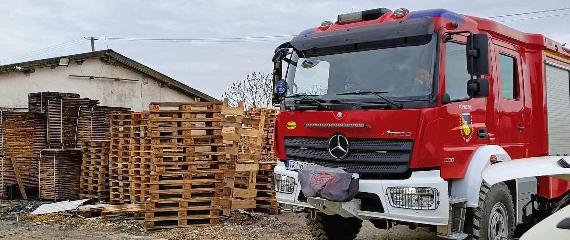 Zdjęcie przedstawia po prawej stronie samochód gaśniczy, a po lewej ustawione palety transportowe na tle hali przemysłowej.