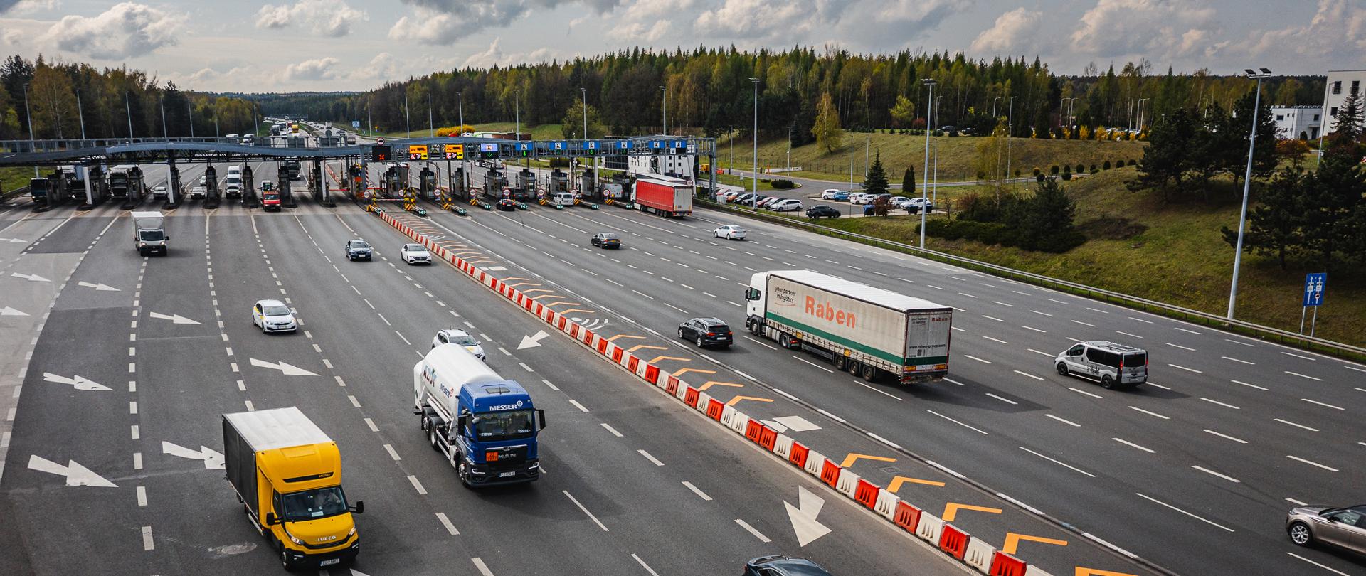Widok z góry na wielopasmowy punkt poboru opłat na autostradzie A4. Przez bramki przejeżdżają samochody osobowe i ciężarowe, a na jezdni widoczne są liczne oznaczenia kierunkowe oraz separatory pasów ruchu. W tle znajduje się zalesiony teren i częściowo zachmurzone niebo. Na zdjęciu widoczna jest infrastruktura autostradowa związana z poborem opłat.