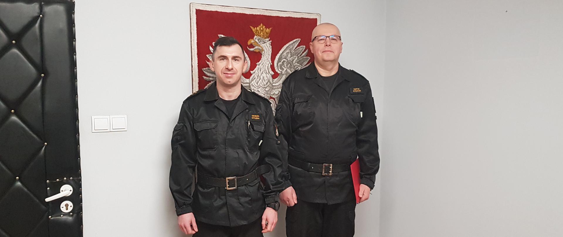 Zdjęcie przedstawia dwóch strażaków - Komendanta Powiatowego PSP oraz dowódcę JRG. Dowódca trzyma w ręce czerwoną teczkę. W tle, na ścianie, znajduje się Godło Polski.