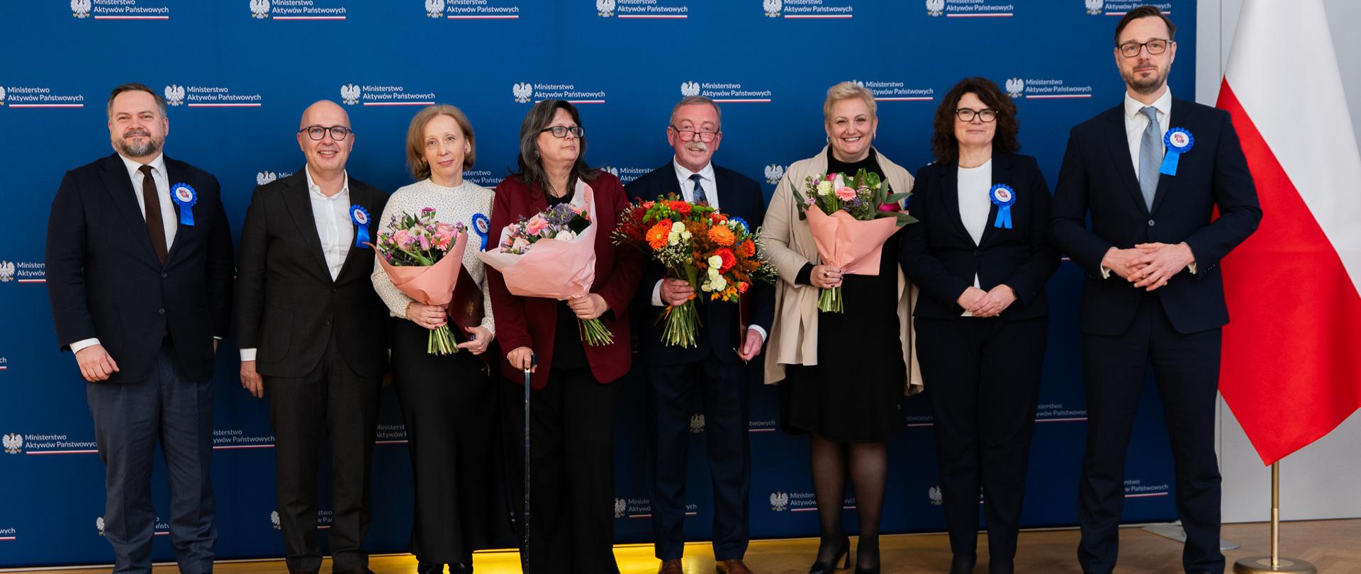 Family photo. Minister Jakub Jaworowski, wiceministrowie: Robert Kropiwnicki i Konrad Gołota, Dyrektor Generalna Ewa Weber oraz pracownicy MAP stoją na tle ścianki z logotypami i flagi. 