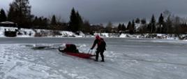 Zdjęcie przedstawia przemyskich strażaków podczas ćwiczenia z zakresu ratownictwa wodno-lodowego , trzech strażaków w skafandrach suchych i kaskach ochronnych w trakcie transportowania strażaka-pozoranta na saniach lodowych