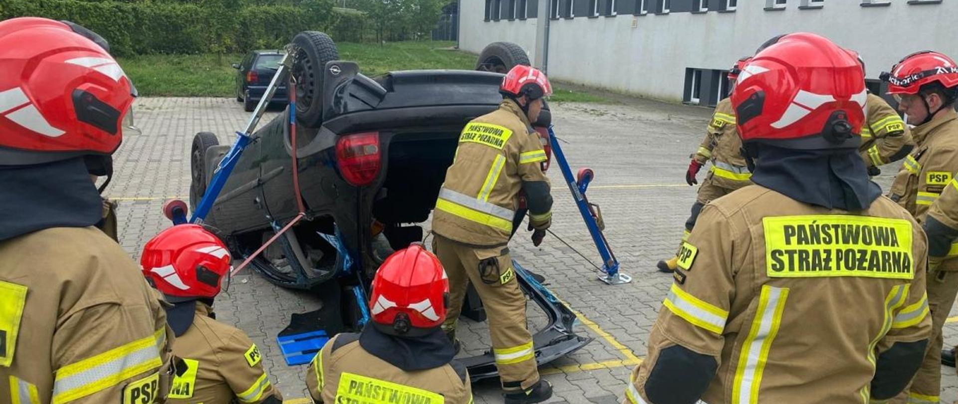 Strażacy Państwowej Straży Pożarnej województwa opolskiego w ubraniach specjalnych i hełmach na głowach uczestniczą w szkoleniu z dziedziny ratownictwa technicznego. Ćwiczenia zorganizowane na placu Komendy Wojewódzkiej Państwowej Straży Pożarnej. Pomiędzy strażakami samochód osobowy leżący na dachu przygotowany do ćwiczeń.