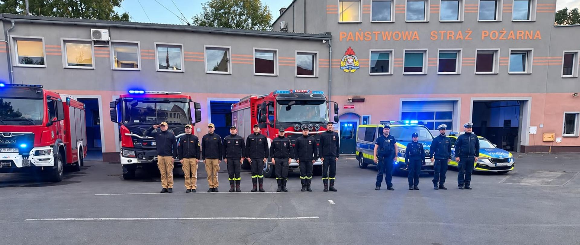 Strażacy i policjanci stoją w szeregu oddając hołd zmarłym strażakom z KP PSP Nisko. W tle pojazdy pożarnicze i radiowozy policyjne z włączonymi sygnałami dźwiękowymi i świetlnymi oraz budynek komendy.