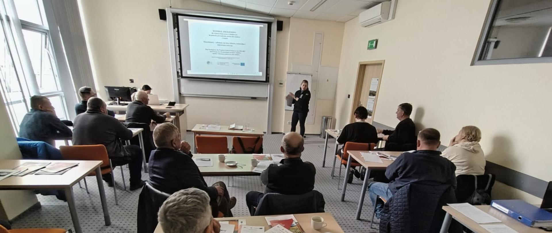 Sala szkoleniowa z kilkunastoma uczestnikami siedzącymi przy stołach, skierowanymi w stronę prowadzącej stojącej przy ekranie projekcyjnym. Na ekranie wyświetlona jest prezentacja. Uczestnicy mają przed sobą materiały szkoleniowe i notatki. Pomieszczenie jest jasne, z oknami po lewej stronie i sprzętem komputerowym przy ścianie z przodu.