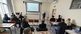 Sala szkoleniowa z kilkunastoma uczestnikami siedzącymi przy stołach, skierowanymi w stronę prowadzącej stojącej przy ekranie projekcyjnym. Na ekranie wyświetlona jest prezentacja. Uczestnicy mają przed sobą materiały szkoleniowe i notatki. Pomieszczenie jest jasne, z oknami po lewej stronie i sprzętem komputerowym przy ścianie z przodu.