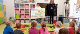 Grupa przedszkolaków siedząca w sali przedszkolnej na kolorowym dywanie w kwiaty. Przed nimi stoją dwie kobiety – jedna z nich trzyma dużą planszę edukacyjną ilustrującą rozwój słonecznika, a druga opowiada. W tle widać regały z różowymi i zielonymi pojemnikami oraz szafki.
