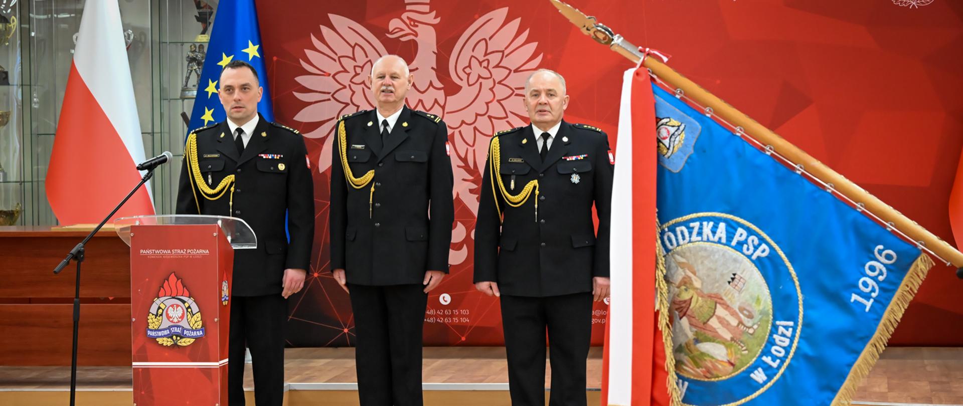 Ujęcie podczas uroczystości: trzech oficerów stoi na baczność w tle, a na pierwszym planie po prawej stronie widoczny jest pochylony sztandar jednostki.