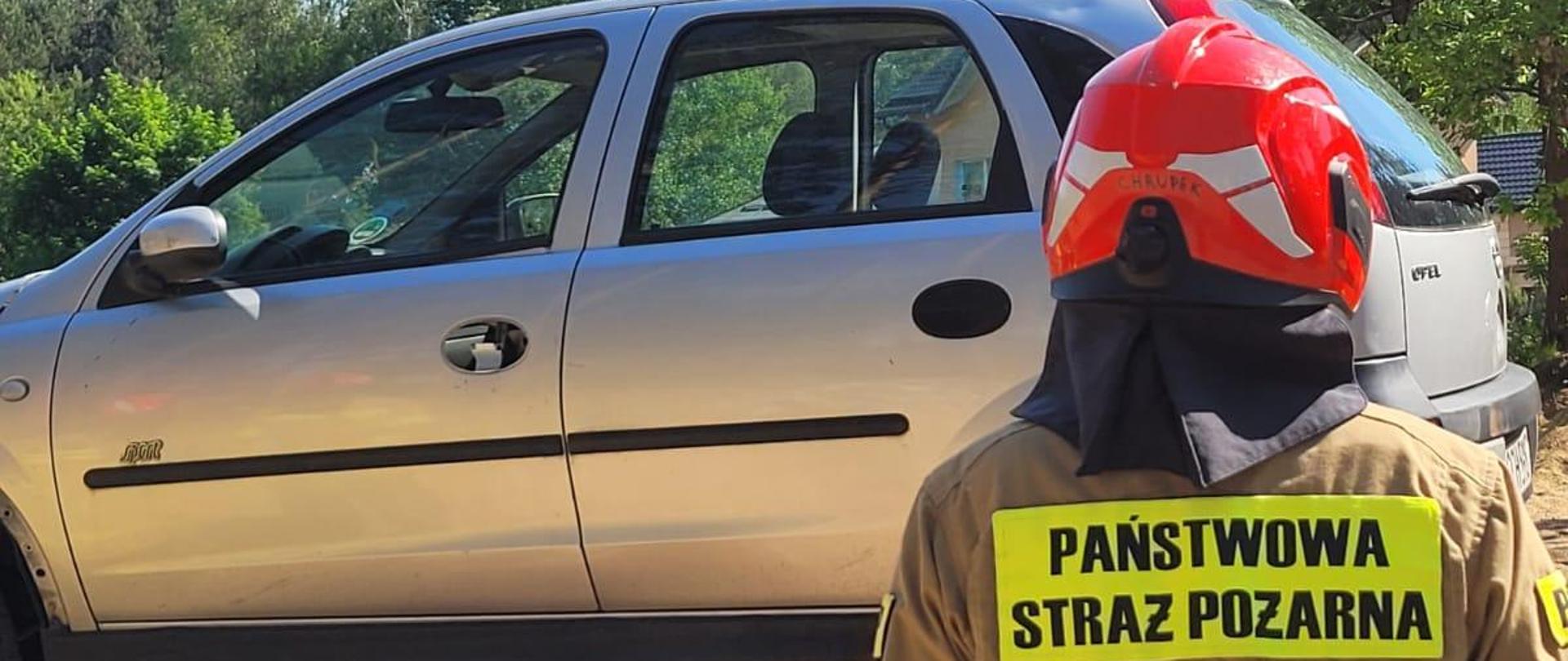 Słoneczny dzień, na tle błękitnego nieba i zielonych koron drzew widać wraki dwóch samochodów osobowych, które są ułożone piętrowo srebrny na dachu czerwonego. Na pierwszym palnie widać plecy strażaka ubranego w piaskowe ubranie specjalne na głowie ma czerwony hełm a na plecach napis dużymi czarnymi literami na jaskrawo-zielonym tle PAŃSTWOWA STRAŻ POŻARNA. 