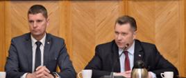 Przy drewnianym stole siedzą minister Czarnek i wiceminister Piontkowski, za nimi ściana wyłożona drewnem, minister mówi do małego mikrofonu stojącego przed nim na stole.