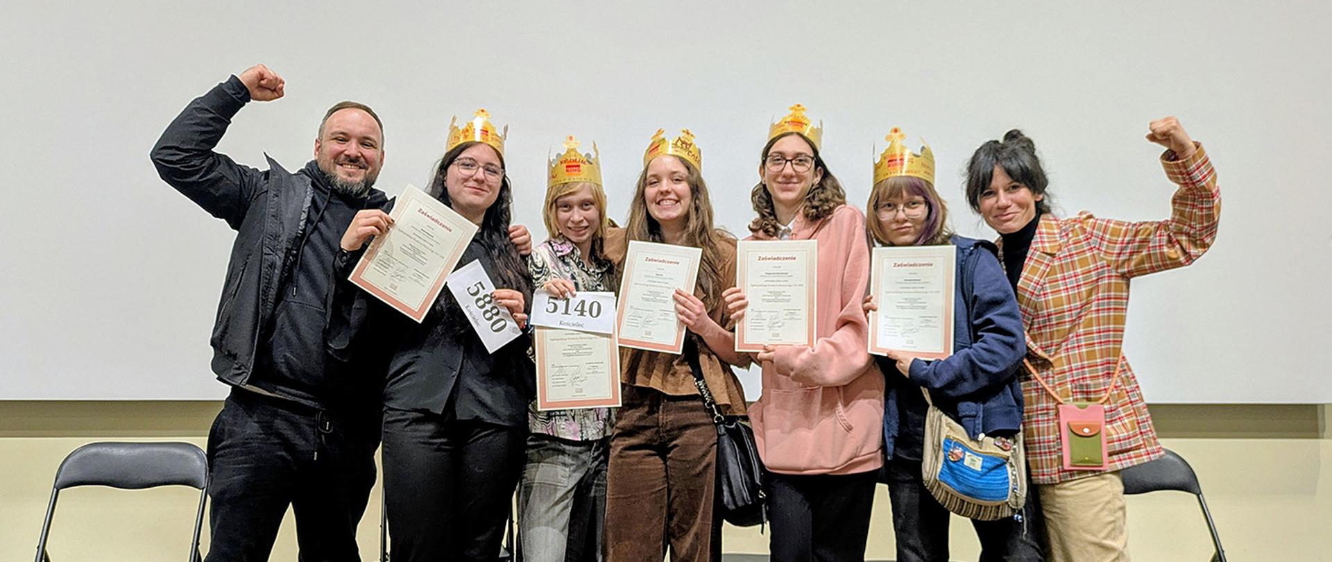 Grupa siedmiu osób stoi na scenie na tle czystej, białej ściany. Po lewej stronie uśmiechnięty mężczyzna napina biceps w geście triumfu. Obok niego pięć młodych uczennic dumnie prezentuje otrzymane dyplomy; każda z nich ma na głowie papierową koronę, co nadaje chwili radosny, swobodny charakter. Po prawej stronie kolejna kobieta również wykonuje gest zwycięstwa. Wszyscy są uśmiechnięci i wyraźnie świętują wspólny sukces.