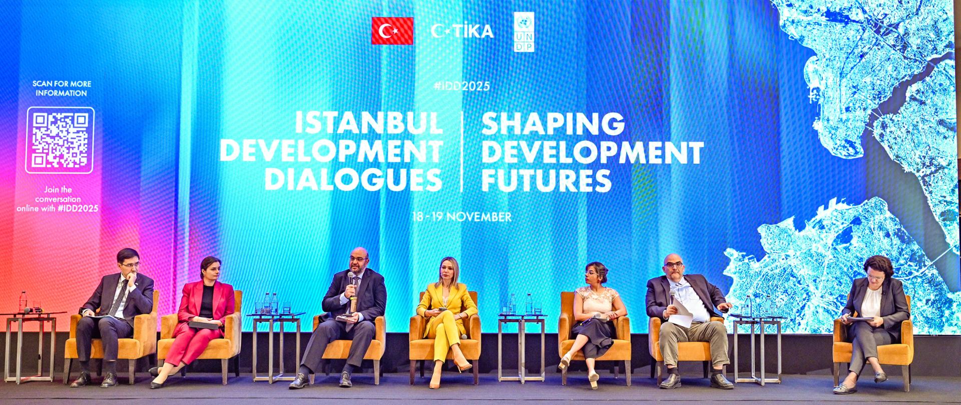 Grupa panelistów siedzi w fotelach na scenie, z tyłu widać napis Istanbul Development Dialogues