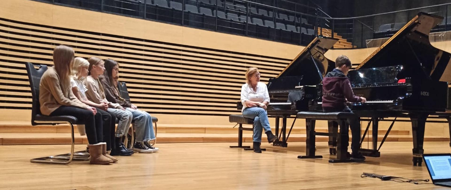 Po prawej stronie estrady sali koncertowej stoją dwa fortepiany, na jednym gra chłopiec, przy drugim siedzi kobieta zwrócona w stronę chłopca. Po lewej stronie na krzesłach ustawionych po skosie w jeden rząd siedzi piątka dzieci.