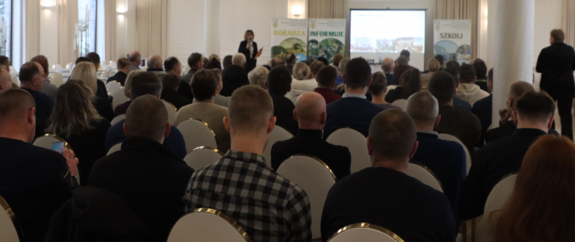 Grupa osób siedzi w sali zwrócona do wykładowcy w głębi; pośrodku prezentacja