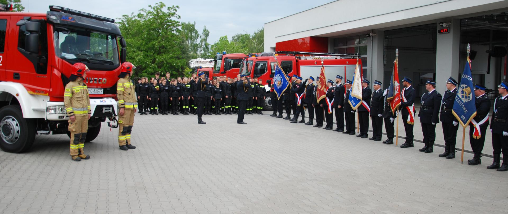 Na zdjęciu po lewej stronie ustawiony jest średni samochód ratowniczo - gaśniczy oraz dwóch strażaków w ubraniu specjalnym podczas robienia przeglądu pododdziałów przez Zastępcę WKW oraz dowódcę uroczystości. 
